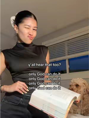 A young woman with dark hair styled with a hair clip, is styled over the shoulder, wearing a black top and holding an open book in her lap. She is winking. A dog is in the background. Text overlays. Warm lighting.