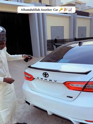 A man in traditional attire stands next to a white car, holding a key fob. The car is parked in front of a building. Overlay text at the top says: " Alhamdulillah Another Car 🔑📈"