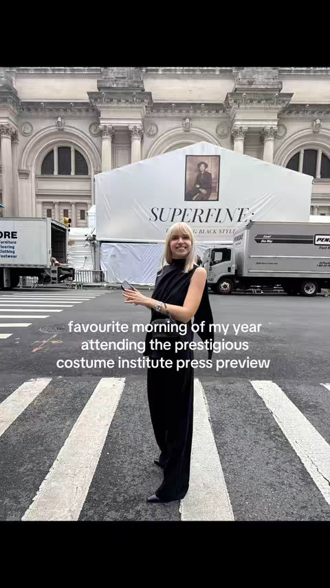 A woman in a black outfit stands on a crosswalk in front of a large building. A sign that says "SUPERFINE" is visible in the background. Below the woman, there is text that says "favourite morning of my year attending the prestigious costume institute press preview".