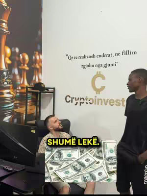 The image shows two men in an office setting. One man is sitting and looking up, while the other stands beside him. The seated man is surrounded by a pile of dollar bills. Behind them, there's a wall with a quote and a logo that says 'CryptoInvest'. On the left side, there's part of a chess set. The frame includes the text overlay 'SHUMË LEKË'.