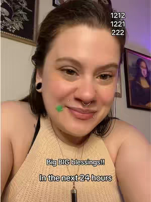 A woman with dark hair poses for a selfie. She has multiple piercings and a green sticker on her face. Text overlays include numbers and a message about blessings. The background includes artwork and a purple-lit wall.