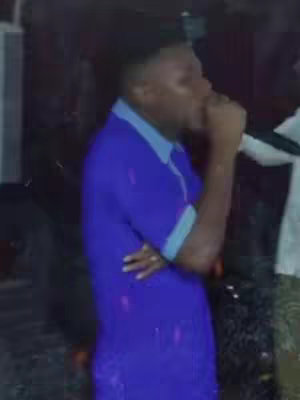 A dark, slightly blurry image of a man speaking into a microphone. The man is wearing a blue collared shirt, and his facial expression suggests he is either singing, preaching, or speaking with great passion. The background is dark and undefined, possibly indoors.