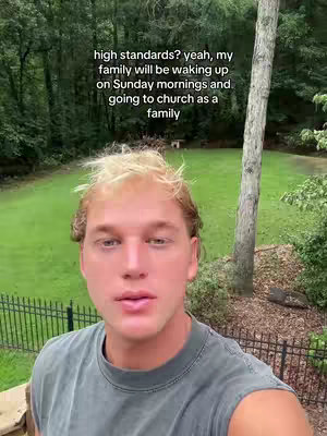 A young man with blonde hair is taking a selfie in an outdoor setting. He's wearing a green t-shirt and the background shows trees, green grass, and a fence.