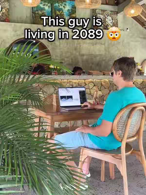 The image shows a man sitting at a wooden table, working on a laptop. The background is a blurred outdoor setting with natural elements and paintings. The text overlay states 'This guy is living in 2089,' with a surprised emoji.