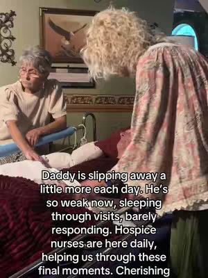 The image shows two women standing near a bed where a man is lying. One woman is sitting by the bed, while the other is standing and leaning over the bed. The room has a painting on the wall and appears to be a bedroom setting.