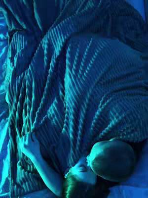 The image shows a person lying in bed, covered by a blue blanket.  The lighting is dim, giving the scene a calming feel. A second person appears to be kissing the lying person, their head and a portion of their shoulder partially visible under the blanket. The overall composition suggests intimacy.