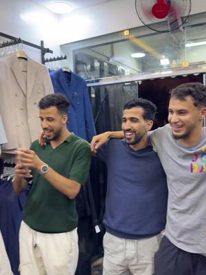 Three young men are smiling and posing for a photo in what appears to be a clothing store. The men stand close to each other, with their arms around each other. The man on the left is holding a phone up, possibly taking a picture. The background shows racks of clothing and a red fan.