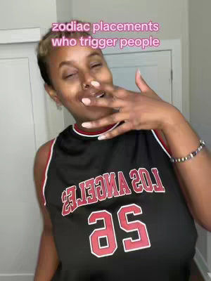 A woman with short dark hair and a nose ring is the central figure. She's wearing a black basketball jersey with red accents and the word "LOS ANGELES" in large red letters. Her right hand is raised towards her face, fingers near her mouth. The background is a plain off-white, suggesting a bathroom setting. The text overlay reads "zodiac placements who trigger people" in a light pink font.