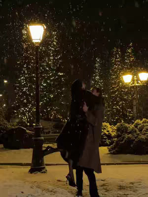 A romantic scene in a snowy evening. A couple is embracing under a lamppost, the man holding the woman in his arms. Snowflakes fill the air against the backdrop of blurred Christmas trees.