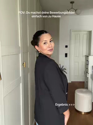 A woman in a black blazer poses in a well-lit room with light-colored walls and a decorative light fixture, looking over her shoulder at the camera. Text overlays indicate this is a POV video, showing how to make application photos at home. In the background, there's a white door, a white cabinet, and a round, upholstered stool.