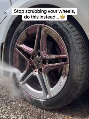 A close-up shot of a car wheel being sprayed with a cleaning solution. The wheel is a silver and gray alloy with the Mercedes-Benz logo in the center. The solution, a dark red color, is being sprayed on the wheel, and the texture of the spray and the wheel is visible.