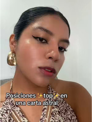 A close-up shot of a woman with smooth skin and dark hair. She has prominent cheekbones, and is wearing large gold hoop earrings. She is wearing makeup including winged eyeliner and dark lipstick. Text overlays the video that reads, "Posiciones top en una carta astral."