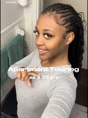 A selfie-style shot of a young woman with long braided hair framing her face. She is wearing a gray long-sleeved shirt and has long eyelashes and earrings. Text overlayed reads 'Apartment Touring as a 23 y/o.' The background is a bathroom setting with a partially visible mirror and a hanging green towel.
