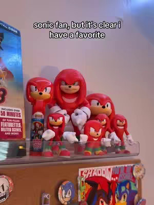 A shelf displays various Knuckles the Echidna figurines. The figures are predominantly red with some touches of white and green. The color scheme is bright and the figures are in various sizes and poses. A box of Sonic movie related items is next to the collection.