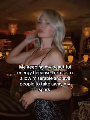A woman with blonde hair and a black dress is standing in a dimly lit room, looking away from the camera. The background is blurred, with visible chandeliers and warm lighting.