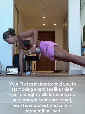 A woman is in a plank position, performing a dumbbell row exercise in a home setting. She wears a sports bra and pink shorts. The background includes a doorway, a laptop displaying a workout video, and parts of a room.