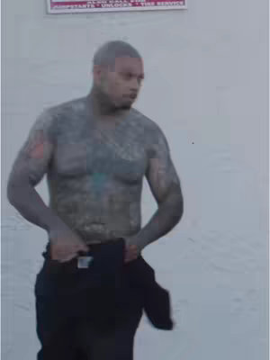 A shirtless man with full-body tattoos adjusts his black pants in front of a mostly white wall. A sign can be partially seen in the background.