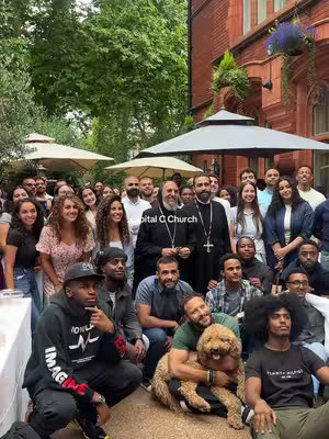 A group of people, men and women, are gathered outside under umbrellas. A few people have crosses on their clothes, and there is a dog involved. The photo is well lit, and it appears to be an outdoor church social. The color palette includes greens, reds, neutrals, and skin tones, with a well balanced composition.