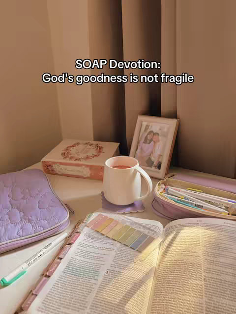 A well-lit indoor scene featuring a desk with a bible open to a reading passage. A cup of tea sits on a coaster, next to the cup is a photo. Other elements include a pencil case, a book, and a journal with a pen. The color palette is soft, with various shades of beige, pink, and lavender.