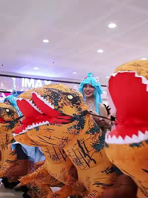 A group of people, some in dinosaur costumes, are gathered in what appears to be a brightly lit indoor space, possibly a mall or event hall. A few people in the image are wearing dinosaur costumes, with the dinosaurs wearing their costumes and open mouths facing forward. The people around the dinosaurs are smiling. The background shows a partial view of a ceiling that could be a mall, along with people behind the dinosaurs.
