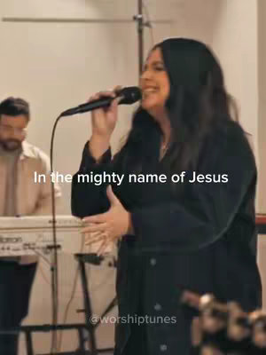 A woman with dark hair is singing into a microphone, and a man in the background is playing the keyboard. The overlaying text is 'In the mighty name of Jesus'.