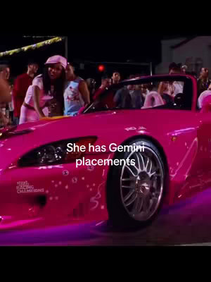 A woman in a pink outfit stands next to a pink car, possibly a sports car, at night with other people in the background. The car has its lights on, and there is text overlayed on the image.