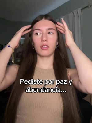 A young woman with long brown hair stands in front of a light-colored background, touching her hair with both hands. Text overlays near the bottom read 'Pediste por paz y abundancia...'