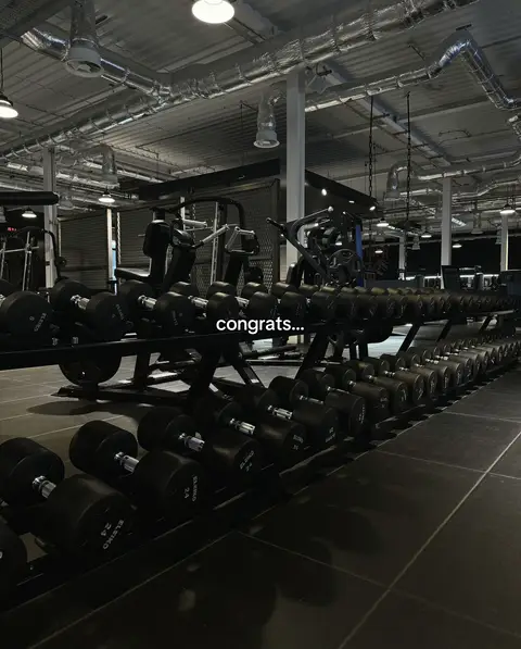 The image is a medium shot taken inside a gym, with a focus on rows of dumbbells arranged on a rack. The rows of dumbbells fill the bottom half of the frame, while the top half gives a glimpse of the gym's ceiling with industrial lighting and ventilation systems. The color palette is predominantly dark, with the metallic dumbbells, racks, and flooring appearing in shades of black and gray.