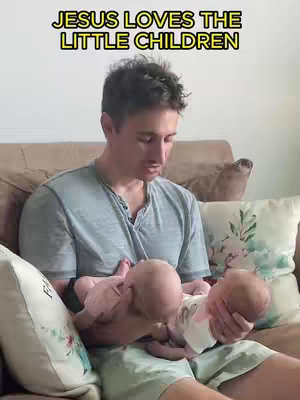 A man sits on a couch, holding two infants. Above the man's head, bright yellow text reads "JESUS LOVES THE LITTLE CHILDREN." The man is looking down, close to the babies. The babies are facing the camera.