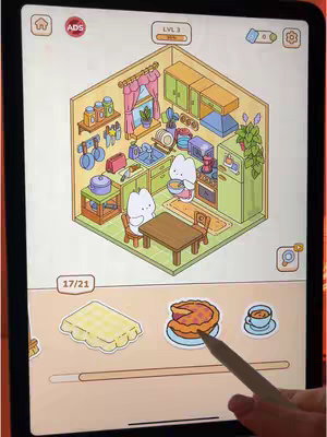 An isometric view of a puzzle game on an iPad. The game shows a decorated kitchen scene with two anthropomorphic cat characters moving around. The bottom of the screen shows puzzle pieces: a waffle, pie, and a cup of coffee.