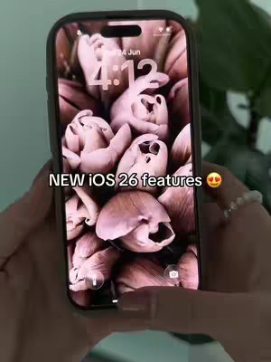 A person holds a phone displaying a pink floral background. The phone has a black frame. The hands holding the phone are visible at the bottom of the frame. Text overlayed on the phone screen says 'NEW iOS 26 features' with a heart-eyes emoji.
