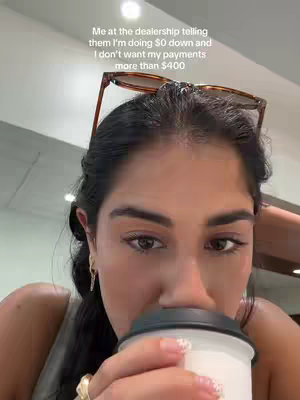 A selfie of a young woman is taken in an indoor setting. She is looking slightly down at a coffee cup she's holding, with the cup obscuring her mouth. She has sunglasses perched on her head and a serious expression. Text is overlaid on the image.