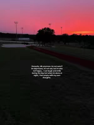 A sunset over a green field and a sports ground. The sky transitions from deep pink at the horizon to a red-orange at the top. A dark silhouette of a tree stands in the middle ground. In the foreground is a green field with the dark outline of the sports track.