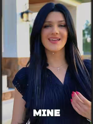 A woman with long black hair wearing a dark top smiles towards the camera in a vertical shot. A gold necklace with a cross is visible.