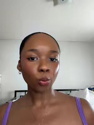 A close-up shot of a woman with smooth, glowing skin. She is puckering her lips. She has dark short hair, and is wearing a purple top and hoop earrings. The background is white with a bed frame visible.