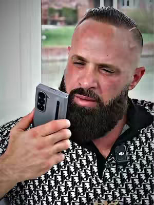 The image features a man holding a phone to his face, looking slightly perturbed. He has slicked-back hair, a full beard, and is wearing a patterned shirt with a small microphone attached. The background is blurred, suggesting the image is tightly focused on the man. The overall composition highlights a moment of communication or phone usage.