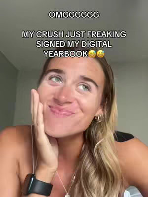 A selfie-style shot featuring a young woman with long blonde hair looking upwards with a happy, slightly surprised expression. The text overlay includes the phrases 'OMGGGGGG' and 'MY CRUSH JUST FREAKING SIGNED MY DIGITAL YEARBOOK'. The background has warm tones, with some interior elements barely visible, highlighting the person in the frame. The lighting is soft and seems natural.