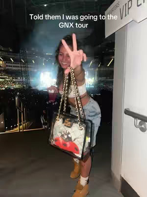 A woman is posing in front of a concert stage, holding a bag and making a peace sign. The background is dark with visible lights and people in the distance.