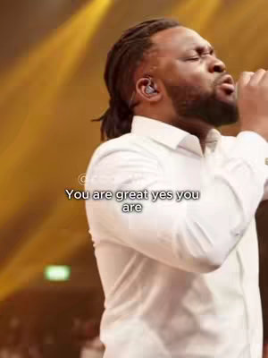 A man with a dark complexion wearing a white button-down shirt is singing into what appears to be a microphone, with his eyes closed and mouth open. The background is blurred, but appears warm-toned, possibly a yellow-orange or gold. Text overlayed reads "You are great yes you are".