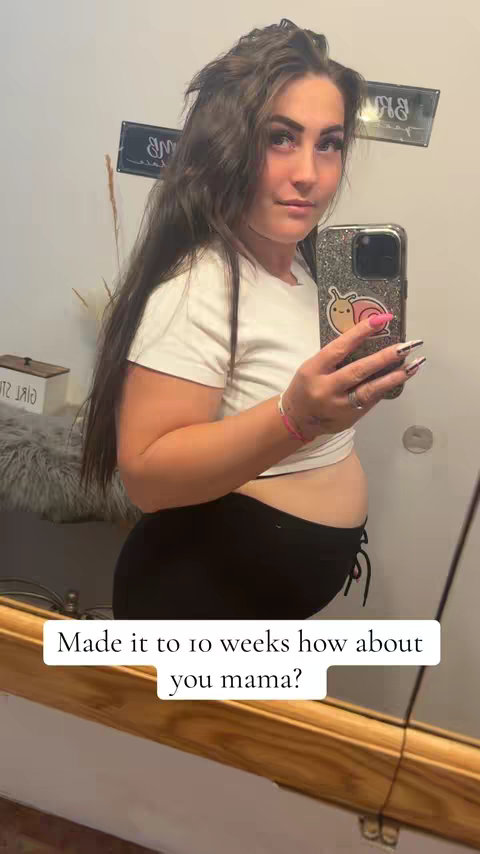 A woman takes a mirror selfie, showcasing her pregnancy. She is wearing a white t-shirt and black leggings. Her hair is long and wavy and her phone has a glittery case. The background includes a bathroom mirror and a sign with some characters on it. The text overlay asks, "Made it to 10 weeks how about you mama?"