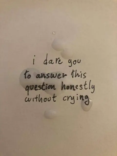 A close-up shot of a clean white surface with text and droplets. The text "i dare you to answer this question honestly without crying" is handwritten in black ink. Irregularly shaped, semi-translucent droplets are scattered around the text, obscuring parts of it. The background is plain and bright.