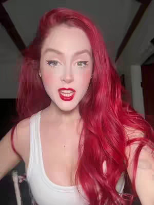 A woman with long, vibrant red hair and red lipstick is looking directly at the camera with an open mouth. She is wearing a white tank top. The background is blurred, showing a room with wooden beams.