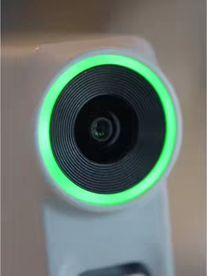 Close-up shot of a camera lens with a bright green ring light. The lens is black with concentric circles, and the outer ring is a vivid neon green. The camera body is a light gray, appearing sleek and modern. The background is blurred, likely an indoor setting. The composition is centered, emphasizing the technological aspect of the camera.