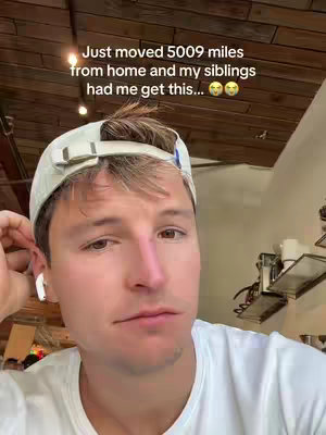 A selfie-style shot featuring a young man with fair skin and light hair wearing a white cap and a white shirt. He appears to be indoors, possibly in a restaurant, with a wooden ceiling in the background. The man is looking at the camera with a somewhat neutral expression, and there is overlay text on the image.