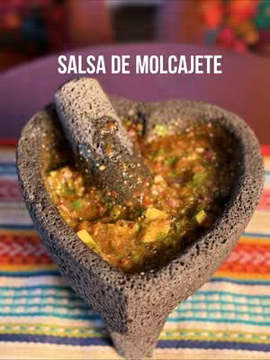 An overhead shot features a heart-shaped molcajete filled with salsa. A pestle rests inside. The salsa is a mixture of colors, greens and reds are present, likely from vegetables. The molcajete sits on a patterned surface of colorful stripes.