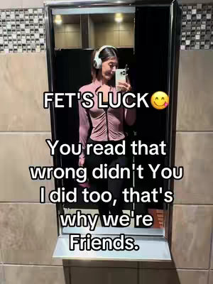 A woman is in a bathroom, taking a mirror selfie. Text overlays say 'FET'S LUCK' and a humorous message about misunderstanding and friendship. She wears a pink top and black pants and has headphones on.