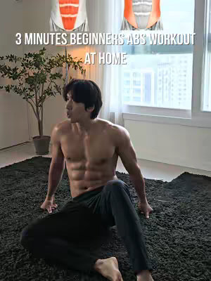 A muscular man is sitting on a dark, shaggy rug in front of a window. He is shirtless, showcasing his defined abdominal muscles. The background includes a small tree in a pot and a view of buildings through the window. The text overlay on the image says "3 MINUTES BEGINNERS ABS WORKOUT AT HOME"