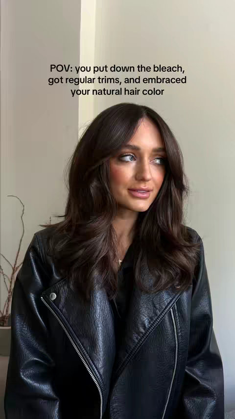 A woman with long, dark brown hair and a black leather jacket looks towards the right of the frame against a neutral-toned background. Text is overlaid on the top.