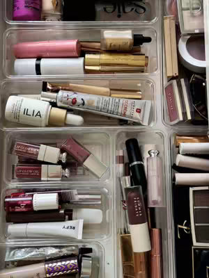 An overhead shot showcasing neatly organized makeup products inside clear plastic drawers. The items are arranged in rows, with lipsticks, lip glosses, foundations, and other cosmetic items visible. The overall composition is clean and well-lit, highlighting the variety of products.