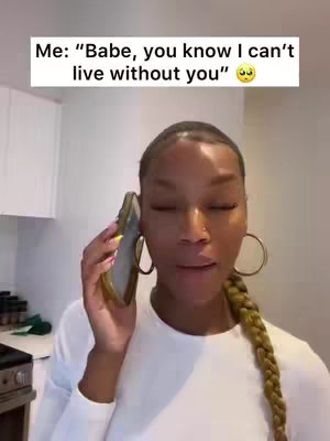 A woman is shown in the kitchen talking on the phone. She is wearing a white top with gold hoop earrings and her hair in a braid. The background includes white cabinets and kitchen appliances.